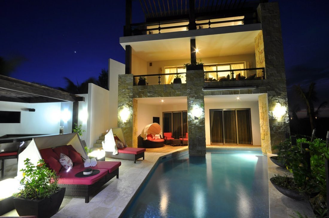 Sereno villa at night with lit terraces on three levels, a stone-clad column, a swimming pool and outdoor lounge beds.